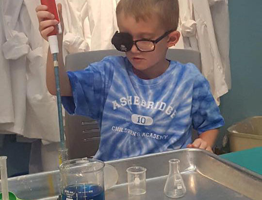 Young preschool boy doing some experiment on colored water in science activity while wearing a special eyeglasses at a Preschool & Daycare/Childcare Center serving Apex & Fuquay-Varina, NC